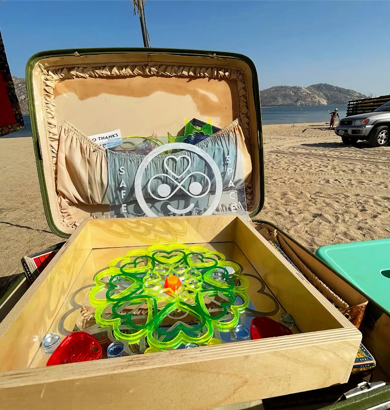 An open vintage suitcase on a beach holding a Safe Space sign and a neon green heart mandala